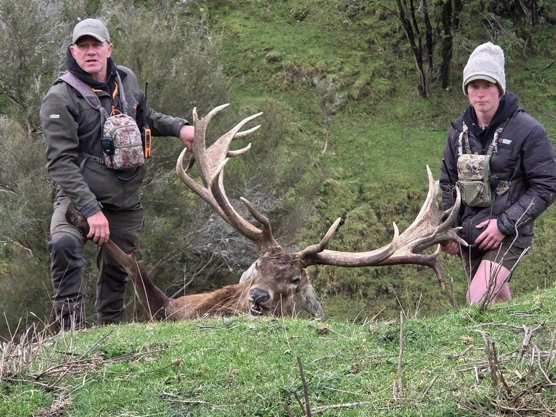 Two hunters with a red stag trophy in a steep green gully